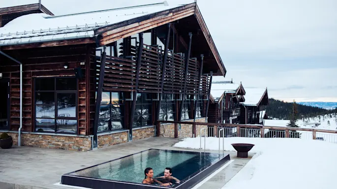 Two people relax in an outdoor heated pool beside a modern wooden lodge. Snow surrounds the area, overlooking a scenic mountain landscape.