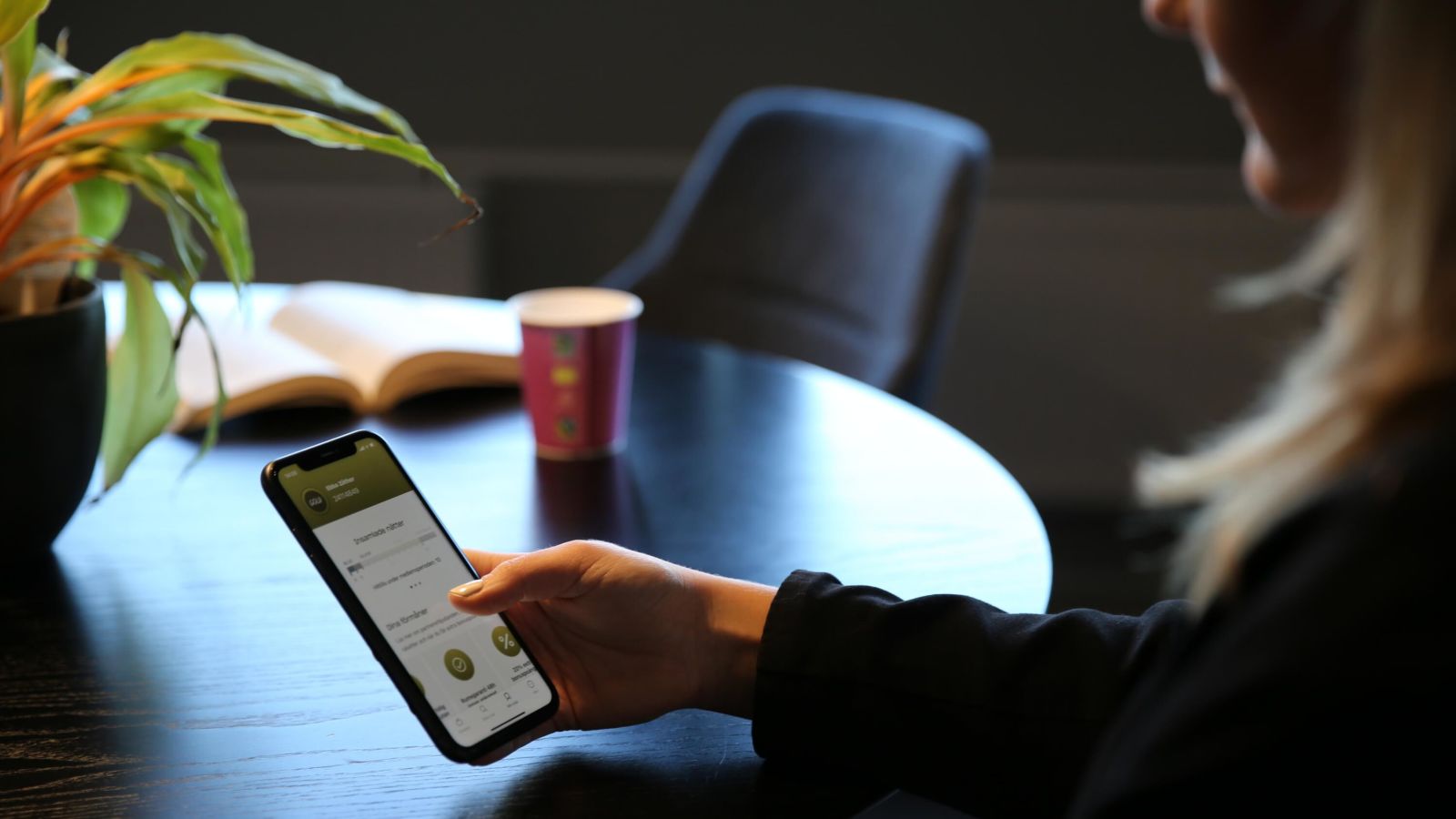 A woman sitting at a table holding up her mobile phone with the Nordic Choice Hotels App.