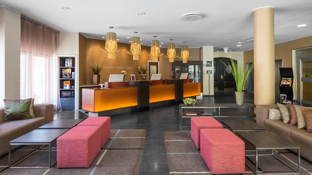 Reception desk with glowing backdrop, displaying "Quality Hotel Luleå" text. Modern lobby features vibrant lighting, plush seating, and decorative plants, creating a welcoming atmosphere.