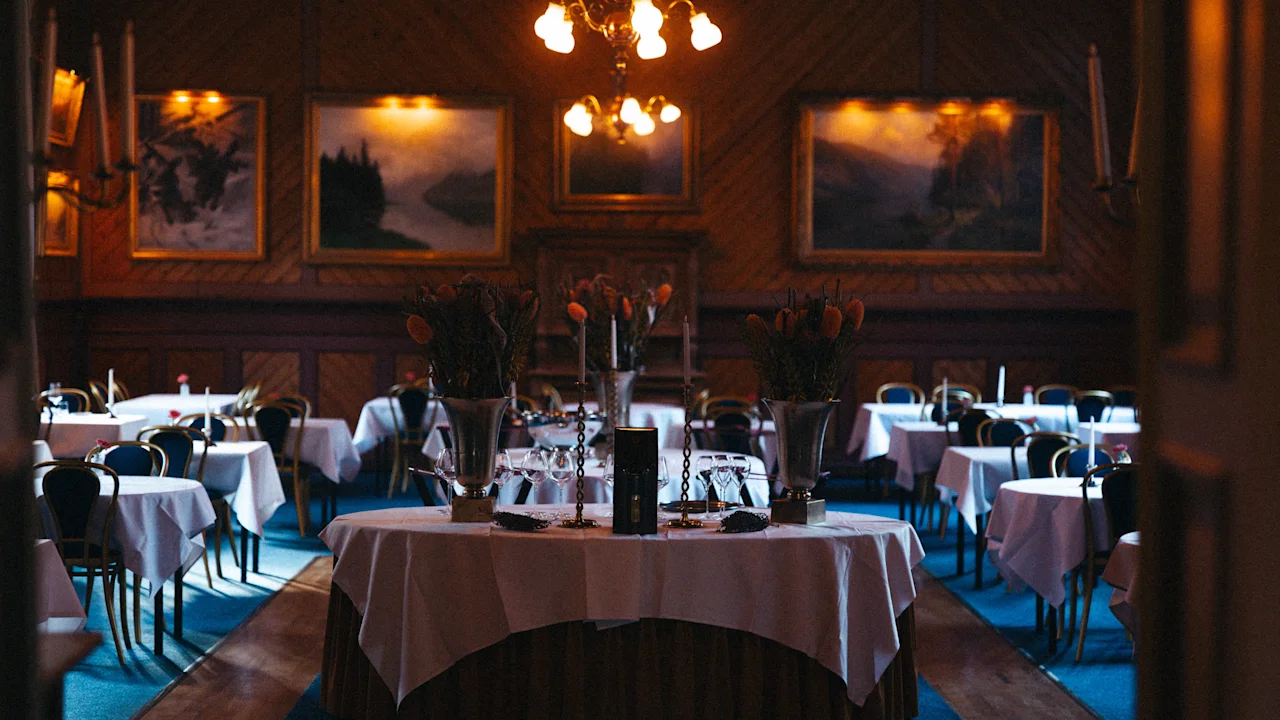 A warmly lit restaurant interior at Dalen Hotel, showcasing elegantly set tables with white linens, soft lighting from chandeliers, and art-lined walls, creating a cozy, inviting atmosphere.