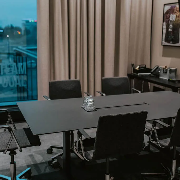 A black conference table with six gray chairs is in a modern office. Behind, a window shows "ONE TEAM ONE GOAL." A side table has a phone. A wall-mounted TV is visible.