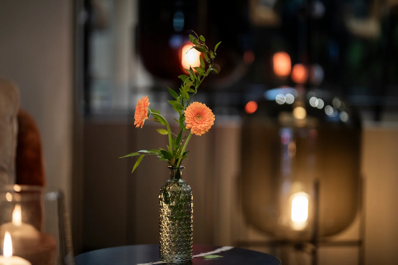 A cozy evening scene at Home Hotel Grand Bodø with a vase of orange flowers on a table and warm, blurred lights in the background.
