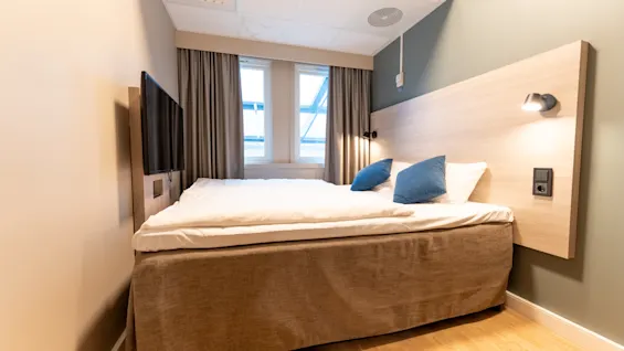 Et moderne hotellrom med en komfortabel seng, blå puter, en sengegavl i tre med lys, en veggmontert TV og vinduer.