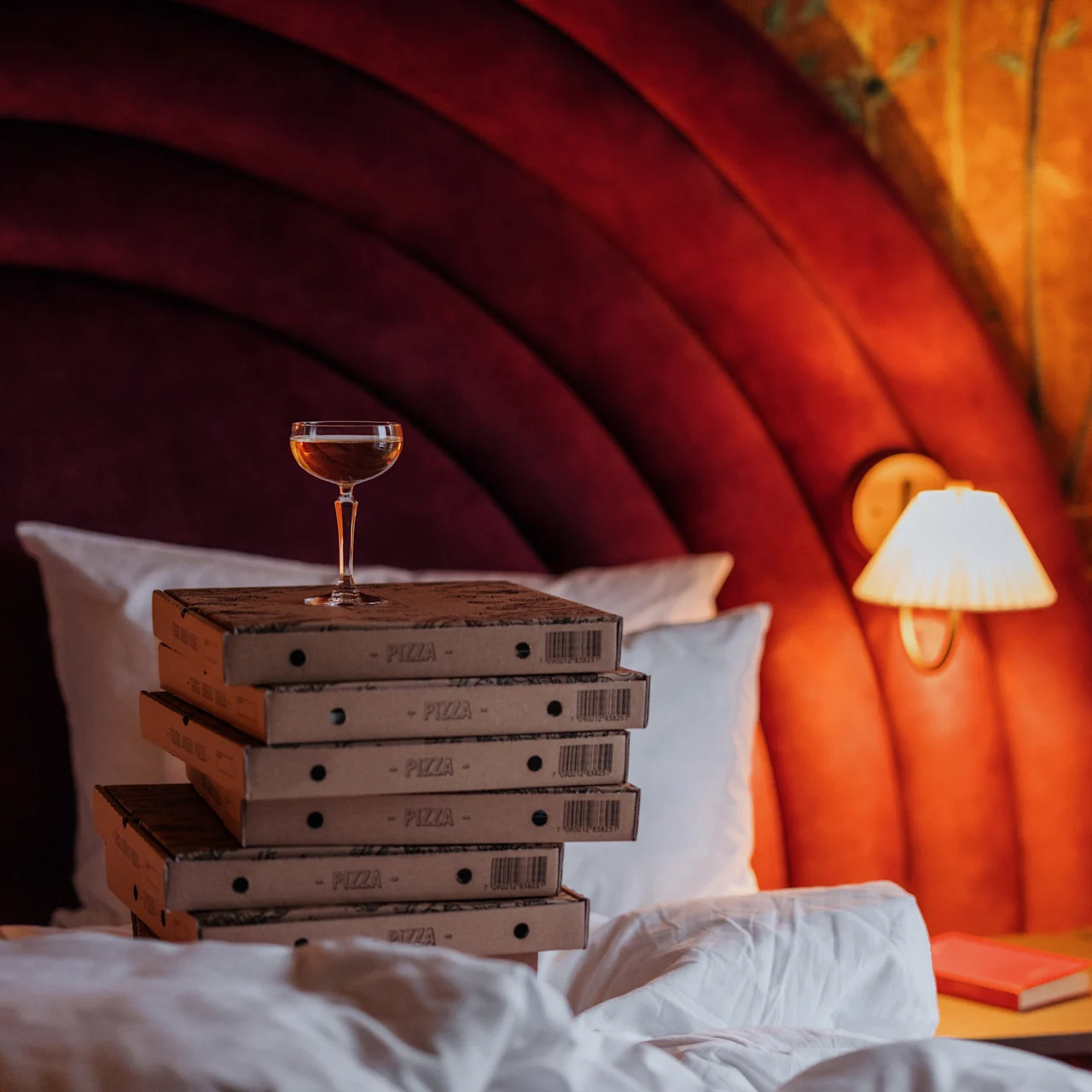 A stack of six pizza boxes supports a glass with a drink, all situated on a messy bed with a red cushioned headboard and soft lamp lighting.