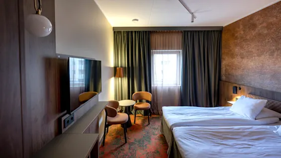 A neatly made double bed beside a wall with a textured finish. Two chairs and a table sit near a curtained window, with a lamp providing ambient light.