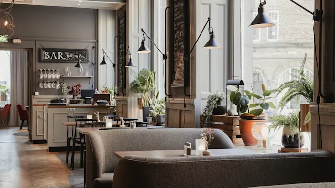 Et varmt og innbydende restaurantinteriør med komfortable båser, bord og en bar. Sollys fyller rommet og fremhever ulike planter. BAR Kvallens Vin.