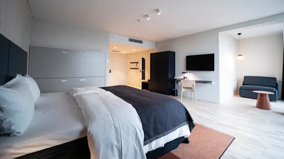 A neatly made bed with dark bedding is in a modern hotel room. Nearby, a wall-mounted TV is above a desk and chair, with a compact sofa and light in the corner.