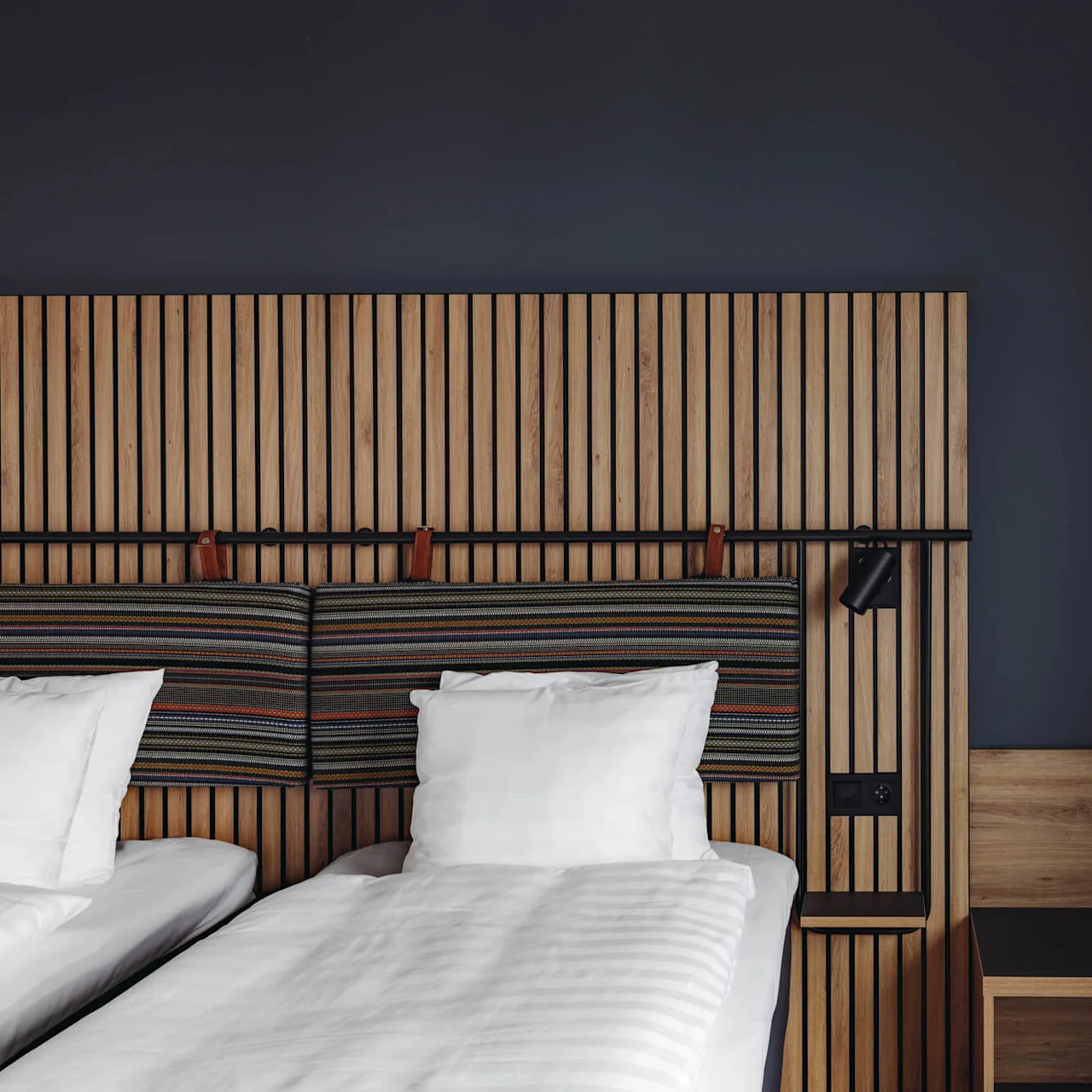 Two neatly made twin beds with white linens stand against a modern wood-panel headboard in a minimalist room, featuring a dark wall and a small wooden nightstand.