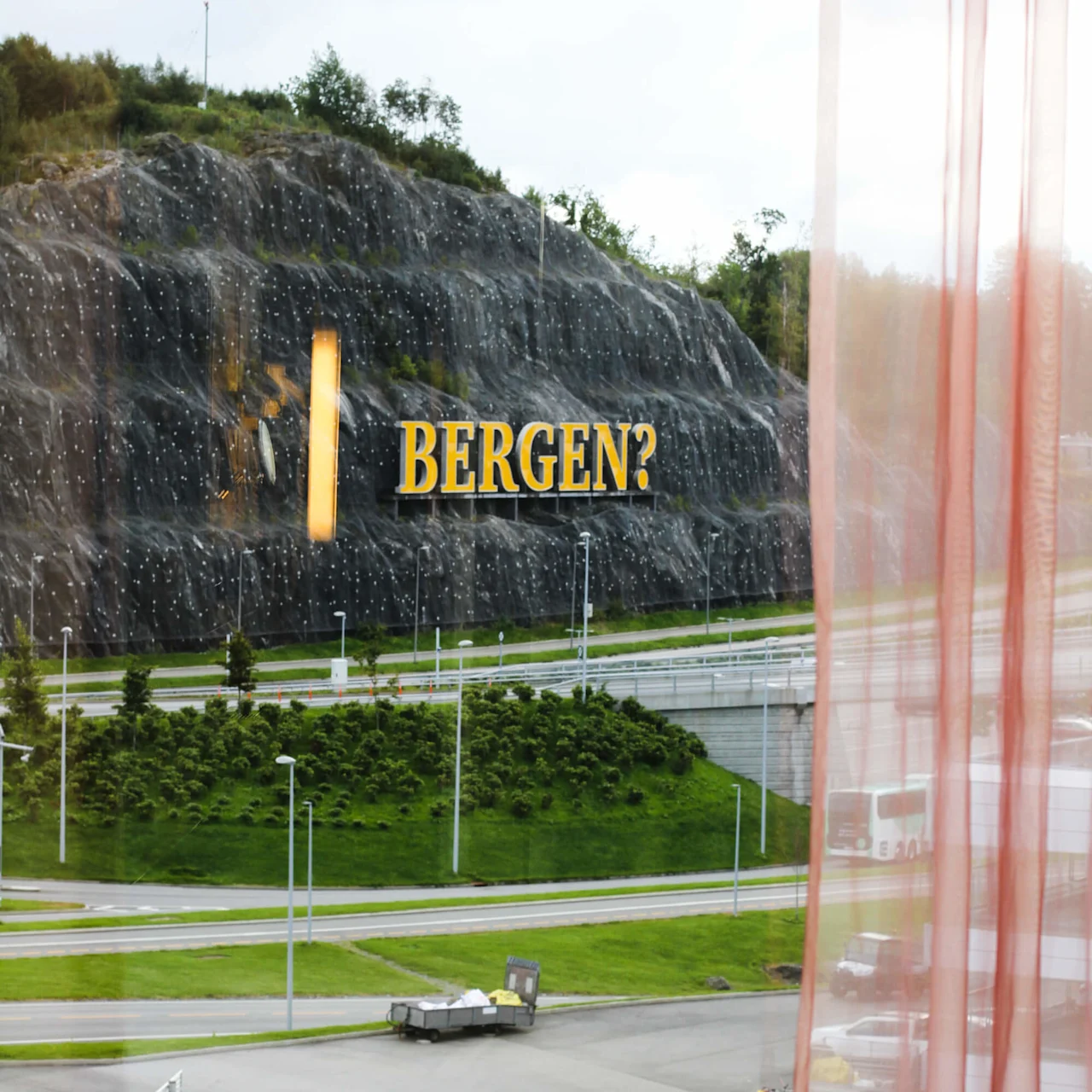 A large yellow sign reads "BERGEN?" on a rocky hillside beside a road. Viewed through a window with sheer curtains.
