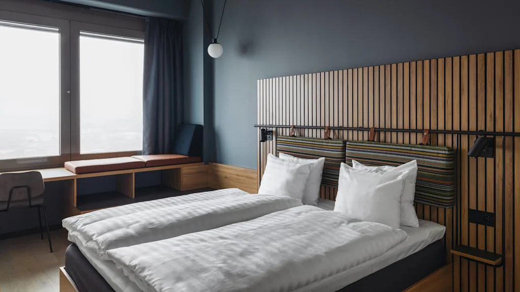 A double bed with white linens is flanked by a wooden headboard with striped cushions. Nearby, a window seat and desk sit by large windows in a modern room.