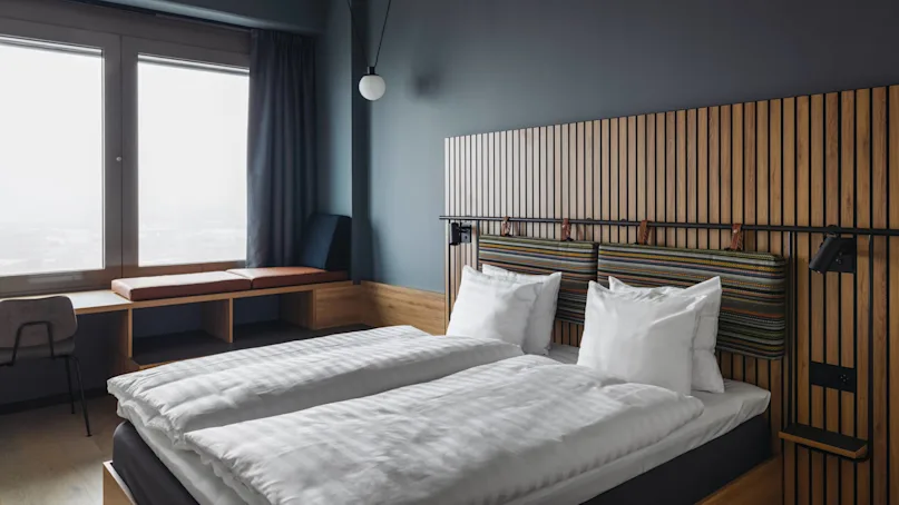 A double bed with white linens is flanked by a wooden headboard with striped cushions. Nearby, a window seat and desk sit by large windows in a modern room.