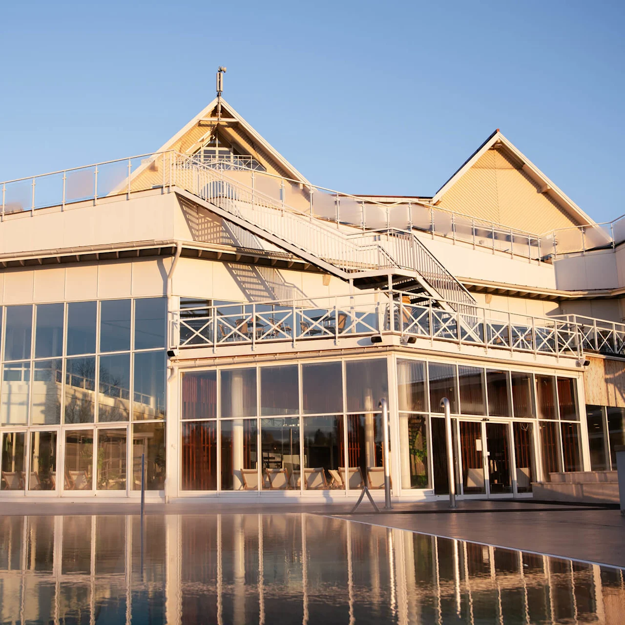 En moderne bygning med store glassvinduer reflekterer sollys, omgitt av en klar blå himmel. En utendørs trapp fører til takområdet og gir arkitektonisk interesse.