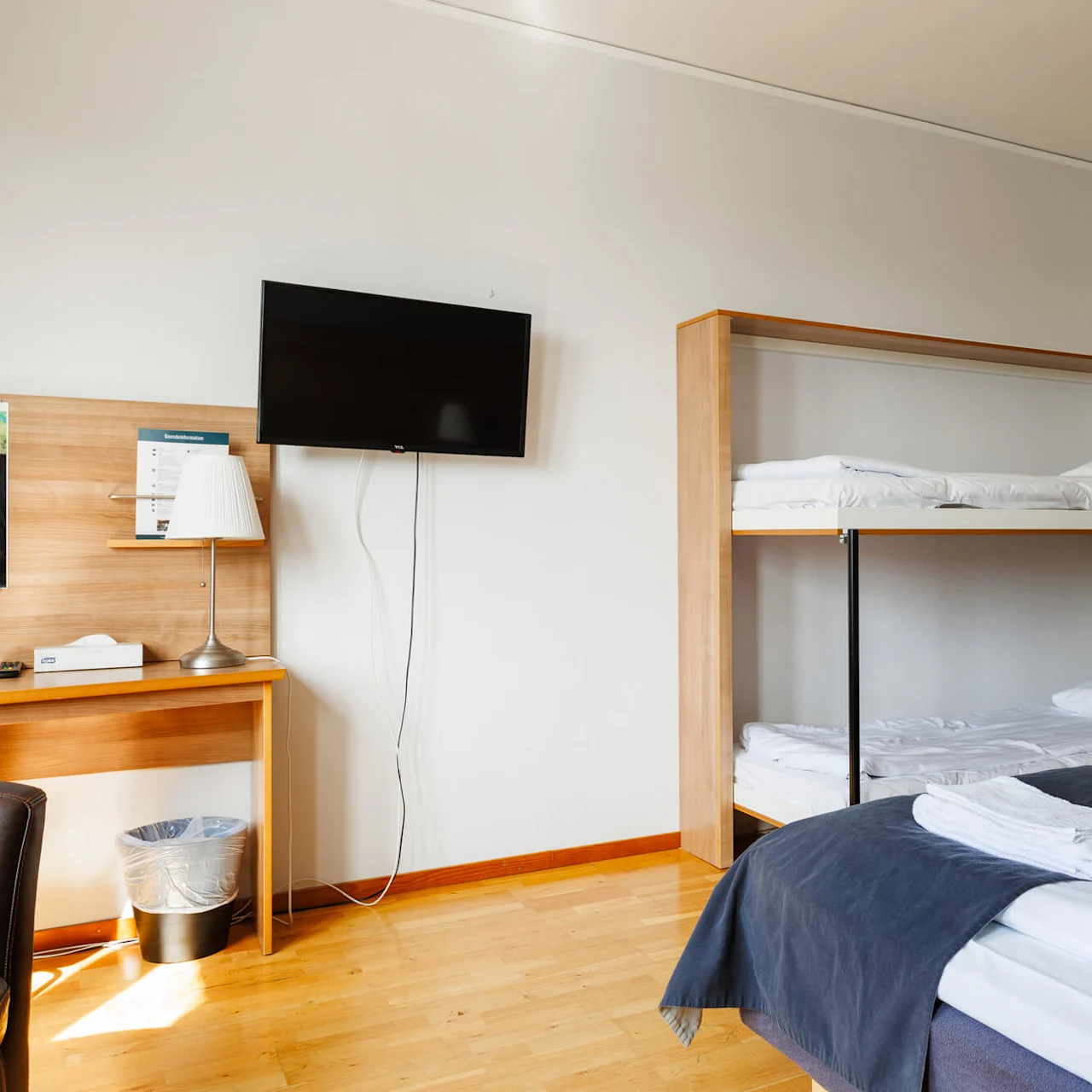 Våningssängar med en trästege står bredvid en prydligt bäddad säng i ett välstädat hotellrum. En väggmonterad TV vetter mot ett skrivbord med lampa och näsdukshållare. 