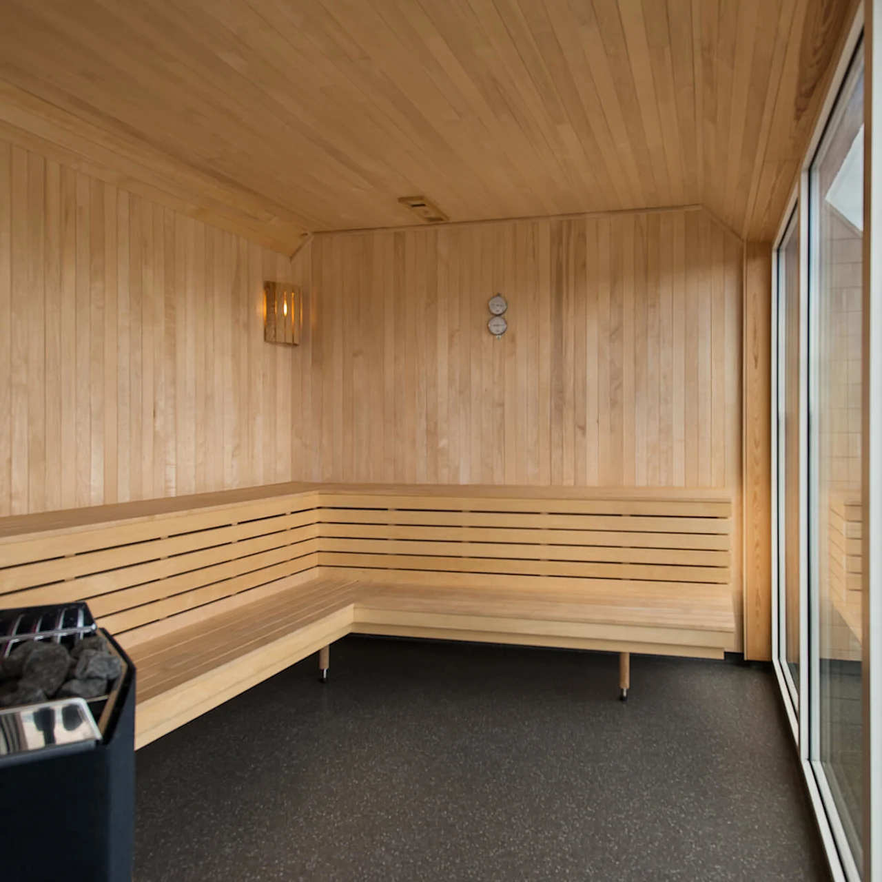 A wooden sauna room with a corner bench, a heater with stones, and a wall-mounted thermometer. Large windows overlook a grassy area with a building visible outside.
