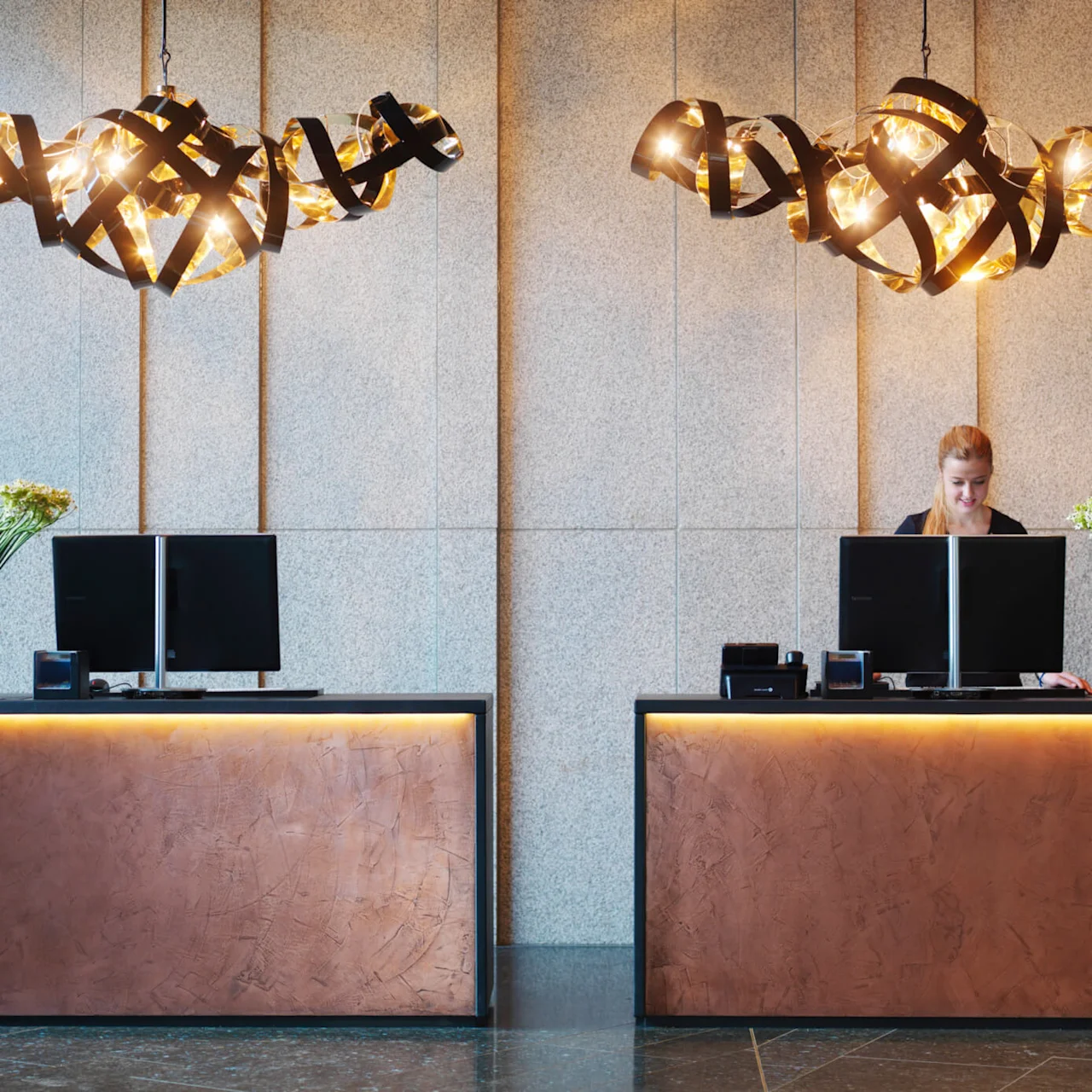 To resepsjonsbord med glødende lamper over, datamaskiner og blomster vaser befinner seg i en moderne lobby. En person står ved ett bord, jobber. Veggen er teksturert og elegant.