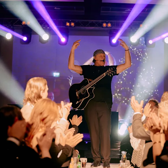 En musiker med gitar opptrer på en lyst opplyst scene, med armene hevet, mens publikum klapper og konfetti faller.