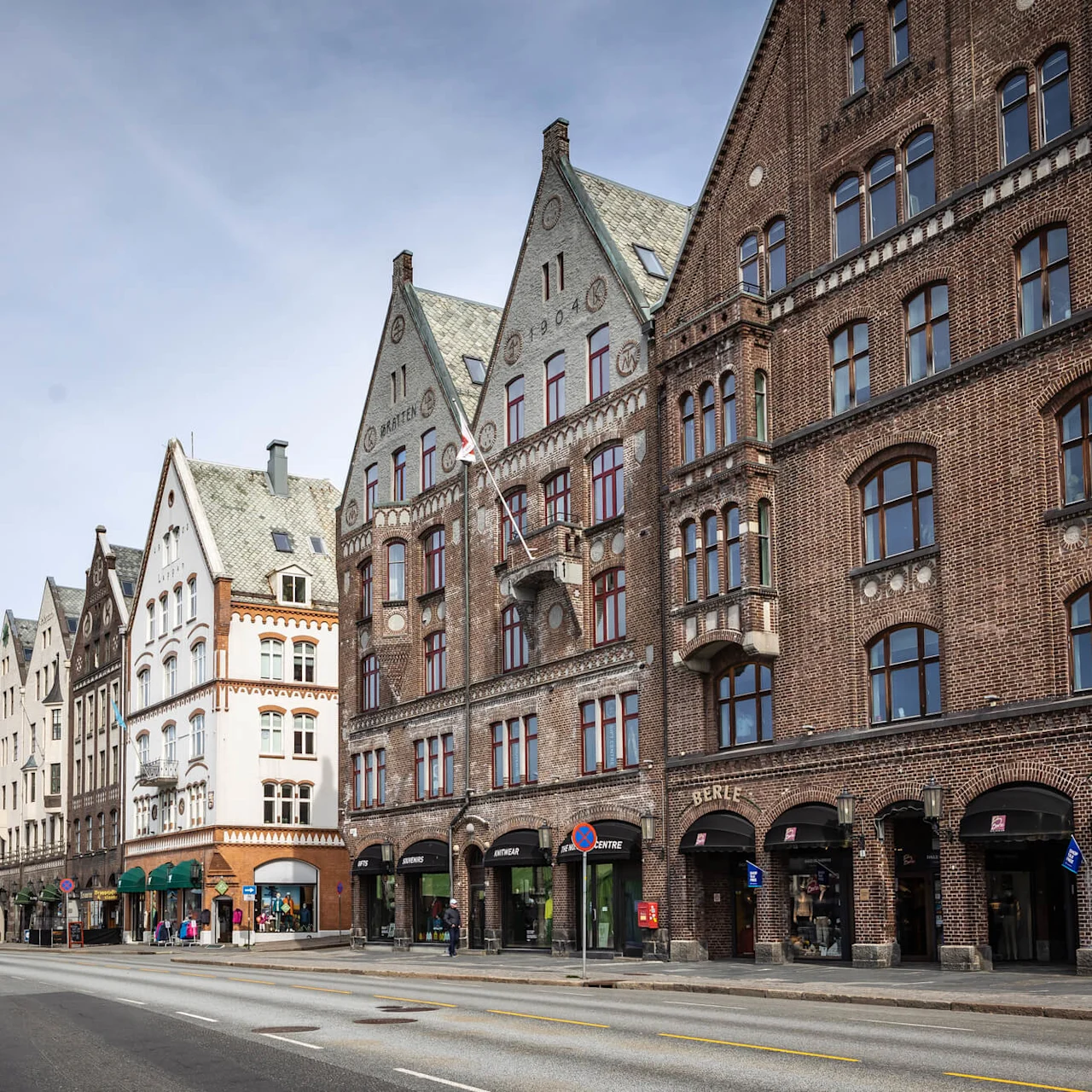 Række af historiske bygninger med gavle af mursten og hvide facader langs en bred, tom gade under en blå himmel. Butiksfacader inkluderer "BERLE" og "SOUVENIRS."