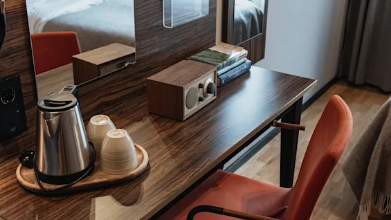 Viihtyisä hotellihuoneen työpöytä, jossa on vedenkeitin, radio ja kirjoja, valmiina työhön tai rentoutumiseen, sängyn heijastuessa peilistä.