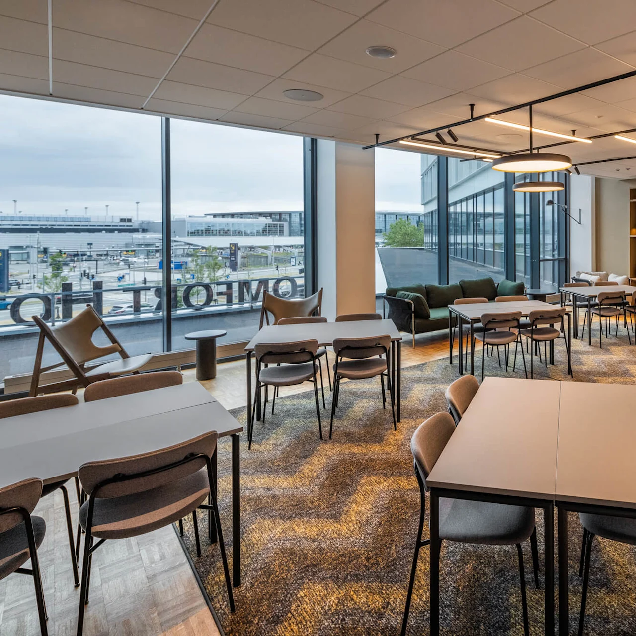 Bord och stolar är arrangerade i ett modernt konferensrum. Stora fönster vetter mot en stadsbild. Det finns bekväma soffor och en stor svart skärm mot en trävägg.