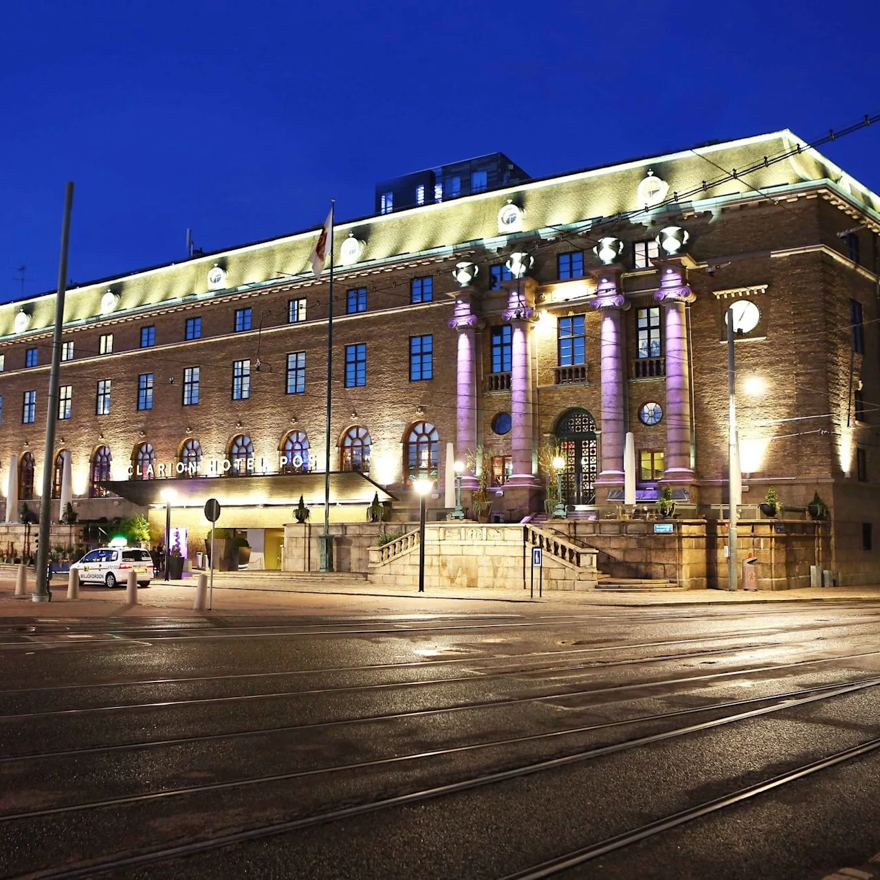 Suuri, valaistu hotellirakennus klassisella arkkitehtuurilla seisoo tummansinistä iltataivasta vasten. Useita pylväitä ja ikkunoita näkyy. "Clarion Hotel Post" on valaistu sisäänkäynnin yläpuolella.