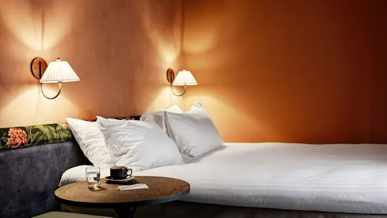Bed with white pillows is beside wall lamps in a softly lit room. A small round wooden table holds a cup, glass, and note card.