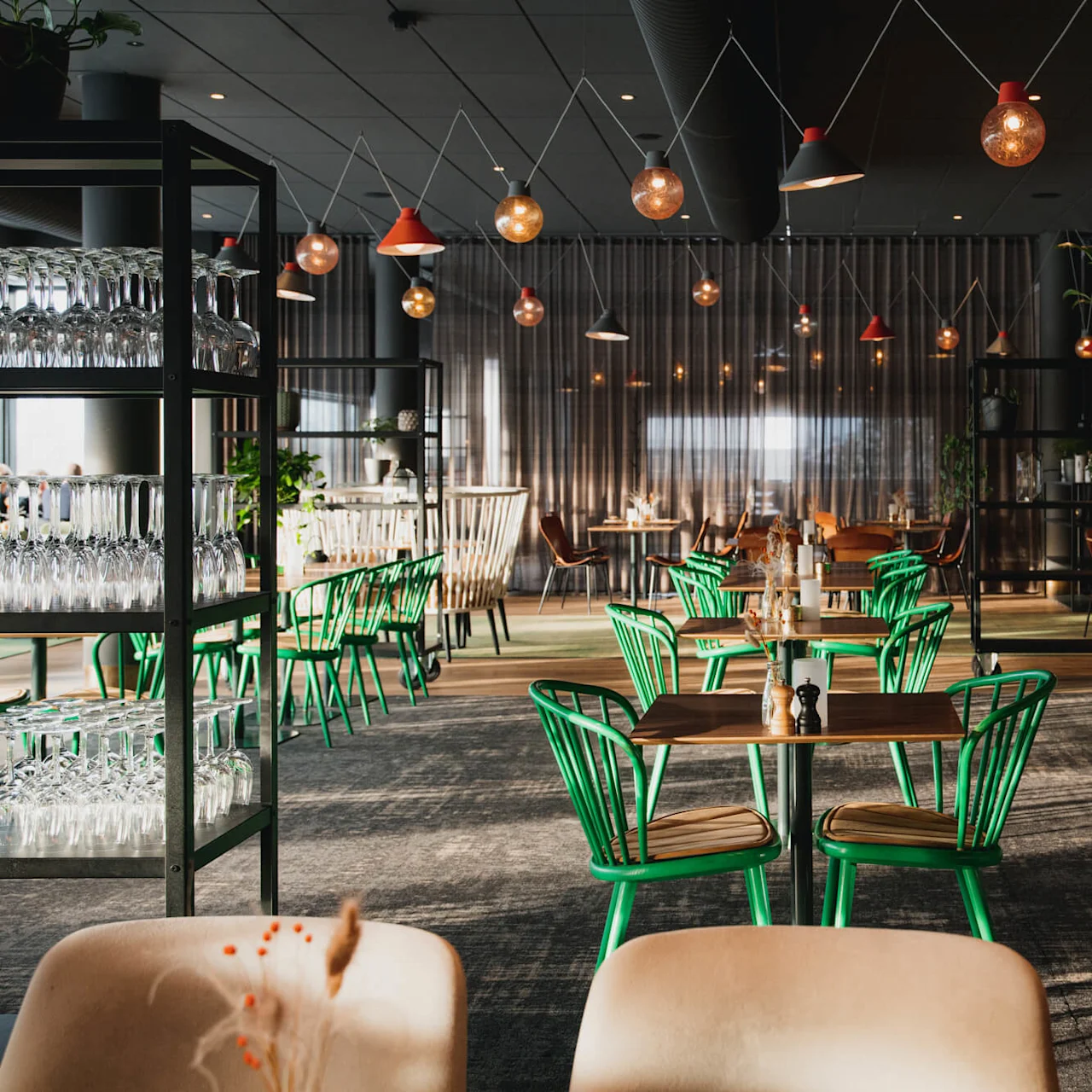 Bord med gröna stolar är placerade, omgivna av hyllor med glas. Hängande lampor finns ovanför i en svagt belyst modern restaurang. En vinställning finns på högra sidan.