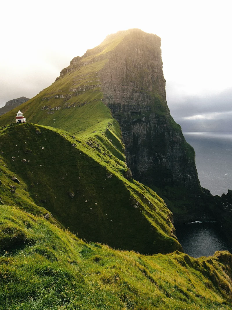 En fantastisk utsikt over en grønn klippe med et lite fyrtårn som skuer ut over havet under en lys himmel på Færøyene.