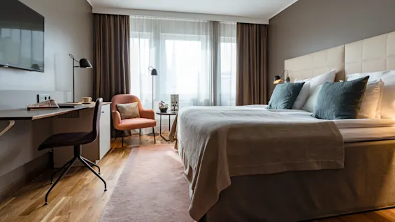 A bed with gray blankets and teal pillows stands in a modern room. A desk with a chair is on the left. Natural light filters through large windows with curtains.