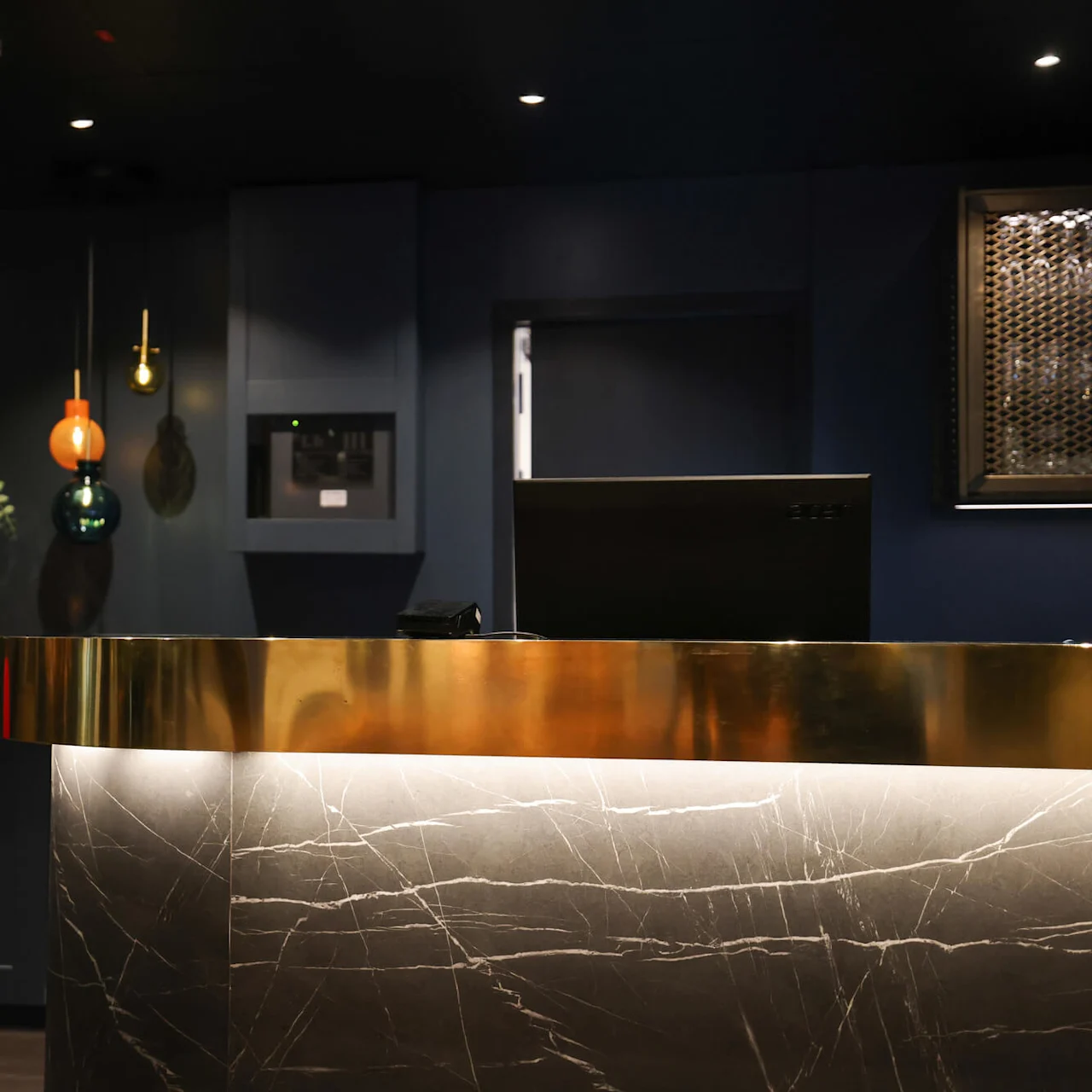 A marble and brass desk stands in a dimly lit reception area; a vase with foliage and modern hanging lights decorate the dark background.