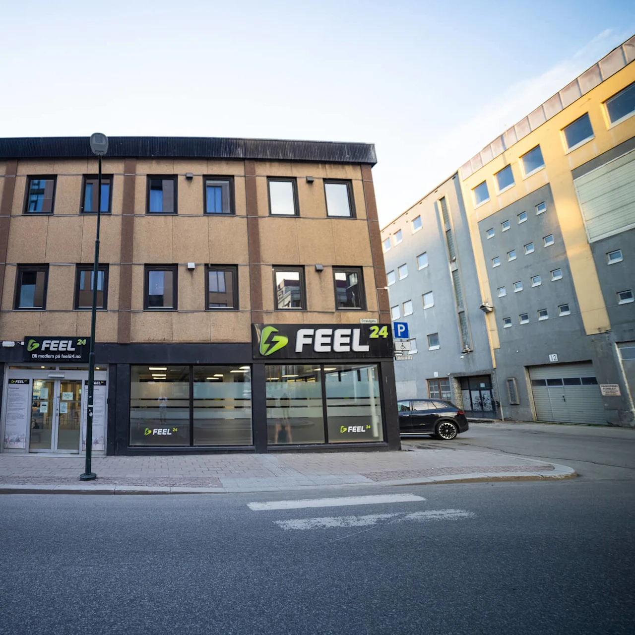 En fitnessbygning viser skiltet "FEEL 24" over indgangen og vinduerne. Det omkringliggende område byder på en gade og tilstødende bygninger. Tekst: "Bli medlem på feel24.no".