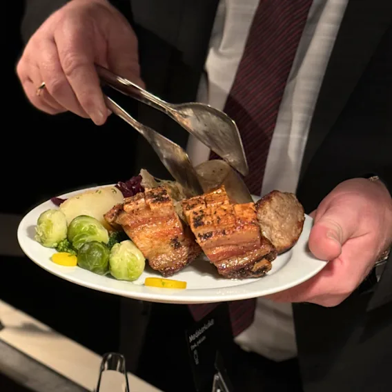 En person serverer et måltid med stekt kjøtt, rosenkål og poteter på en hvit tallerken med tang.