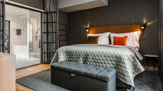 En seng med en tufted sengegavl står op ad en mørk væg, med grå og orange puder. Den vender mod et glasvægget bad i et moderne, varmt belyst værelse.