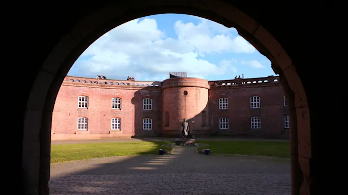An archway frames a courtyard with a statue centered on cobblestone ground. The surrounding red brick building features multiple windows. Blue sky and clouds are visible overhead.