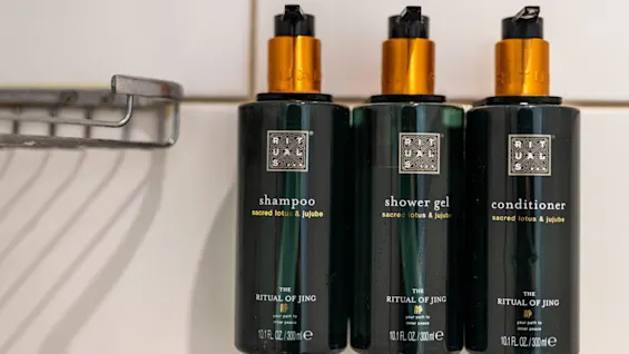 Schampo, duschgel och balsamflaskor står sida vid sida på en hylla i badrummet. Text på varje lyder: "sacred lotus & jujube, THE RITUAL OF JING, your path to inner peace, 10.1 FL OZ / 300 ml."