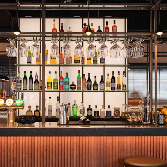 Barbord med olika spritflaskor på hyllor, omgivna av hängande glas. Tappar för fatöl syns i en rymlig, modern bardmiljö.