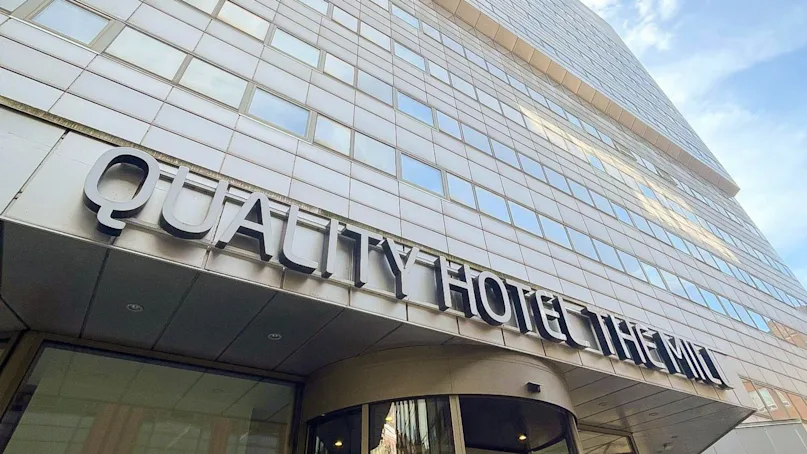 Quality Hotel The Mill sign adorns a modern building's facade, stretching above a glass entrance with revolving doors, under a blue sky.