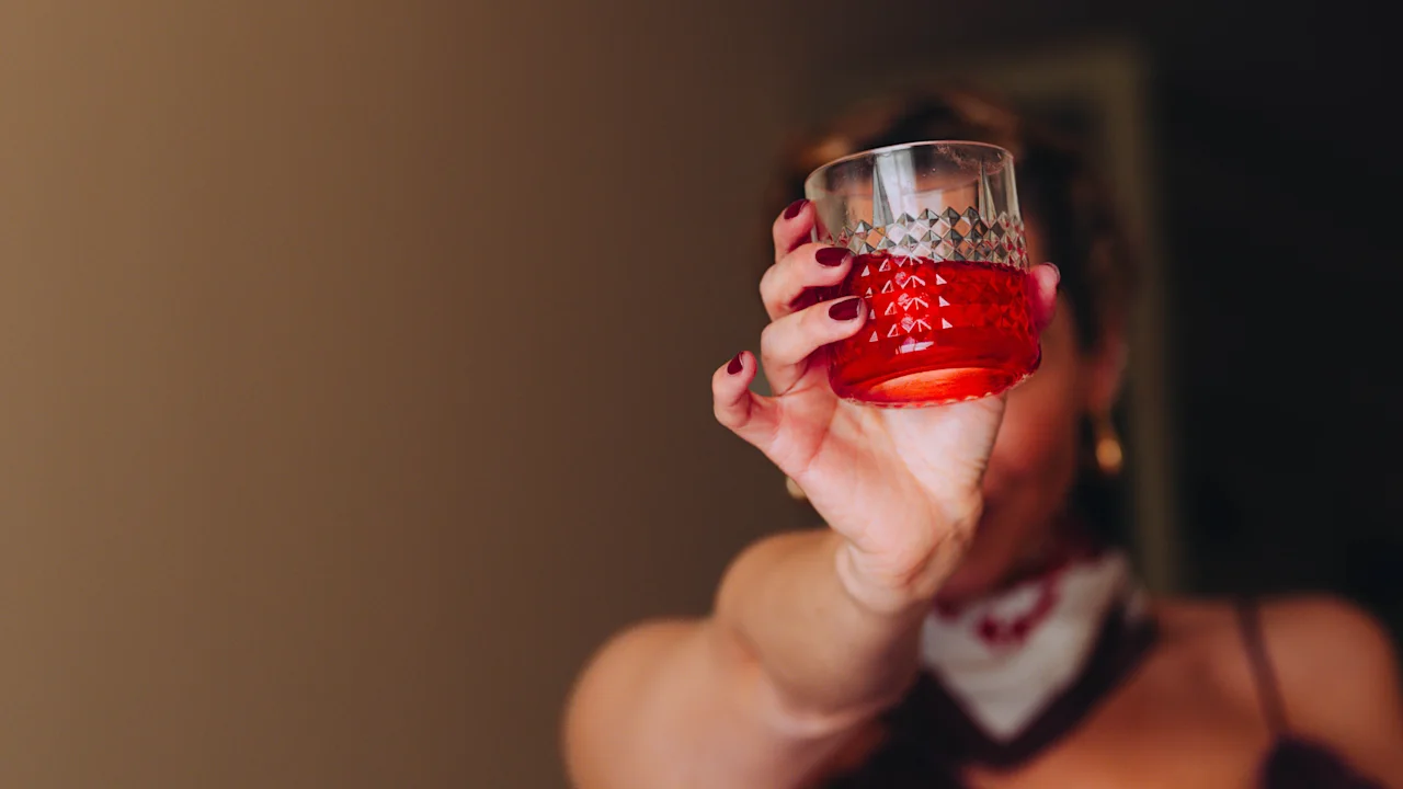 En kvinna i en lila klänning håller en röd drink i ett glas, armen utsträckt mot kameran, inomhus med dämpad belysning.
