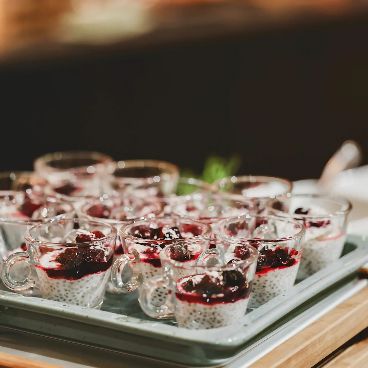 Small glass cups hold chia pudding topped with dark berry sauce, arranged neatly on a tray, all placed on a wooden table in an indoor setting.