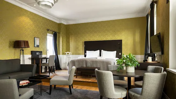 A spacious hotel room features a bed with white linens, seating area, and dining table. Green wallpaper, dark curtains, and a flat-screen TV provide a stylish and cozy atmosphere.