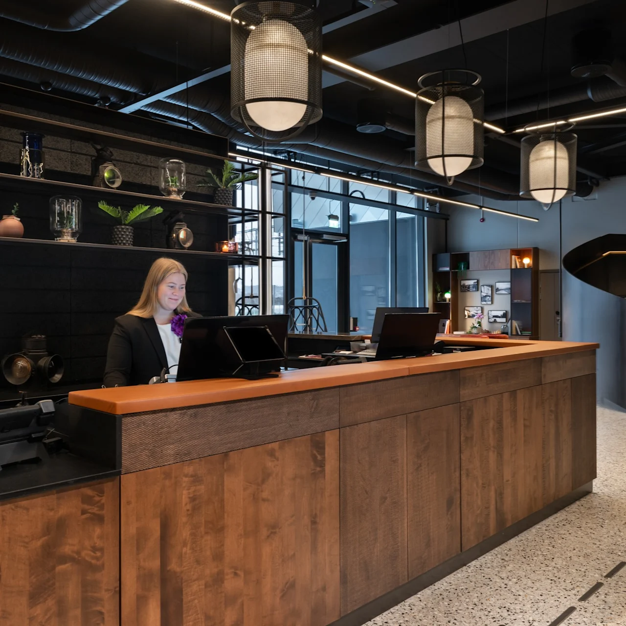 Resepsjonist på jobb ved en moderne hotellresepsjon med trepanel; omgitt av dekorative hyller, lysarmaturer og en vindeltrapp som leder oppover i en velbelyst, åpen plass.