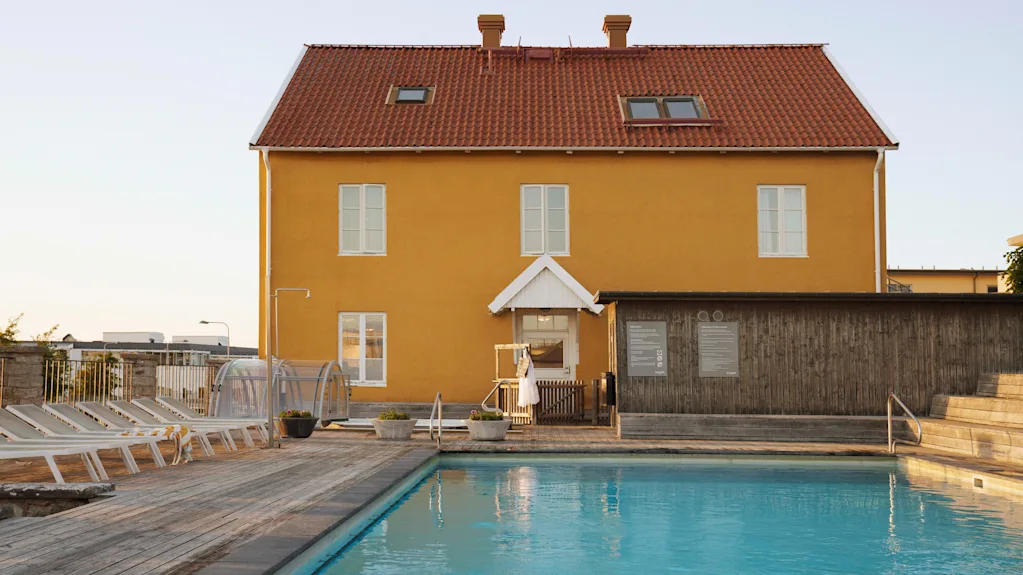 En livlig gul byggnad med rött tak vetter mot en uppfriskande utomhuspool med solstolar en solig dag.