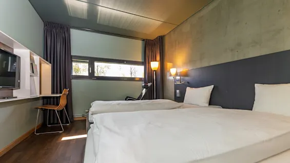 Two neatly made beds rest in a modern hotel room with wooden floors. A desk with a chair sits by a window with curtains, illuminated by wall-mounted lamps.