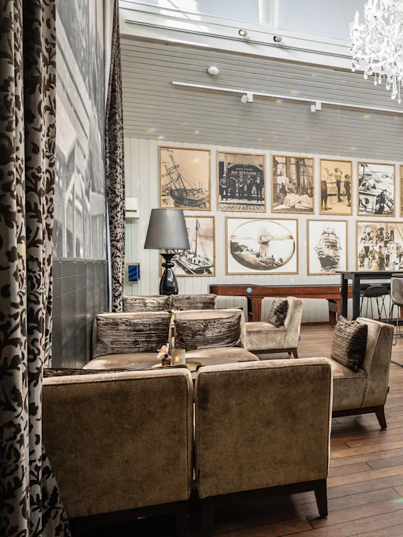 En lyxig ljuskrona hänger ovanför en elegant lounge med vintagefotografier på väggarna, bruna stoppade stolar runt borden. Ljust naturligt ljus filtreras genom stora fönster och skapar en sofistikerad, luftig atmosfär.