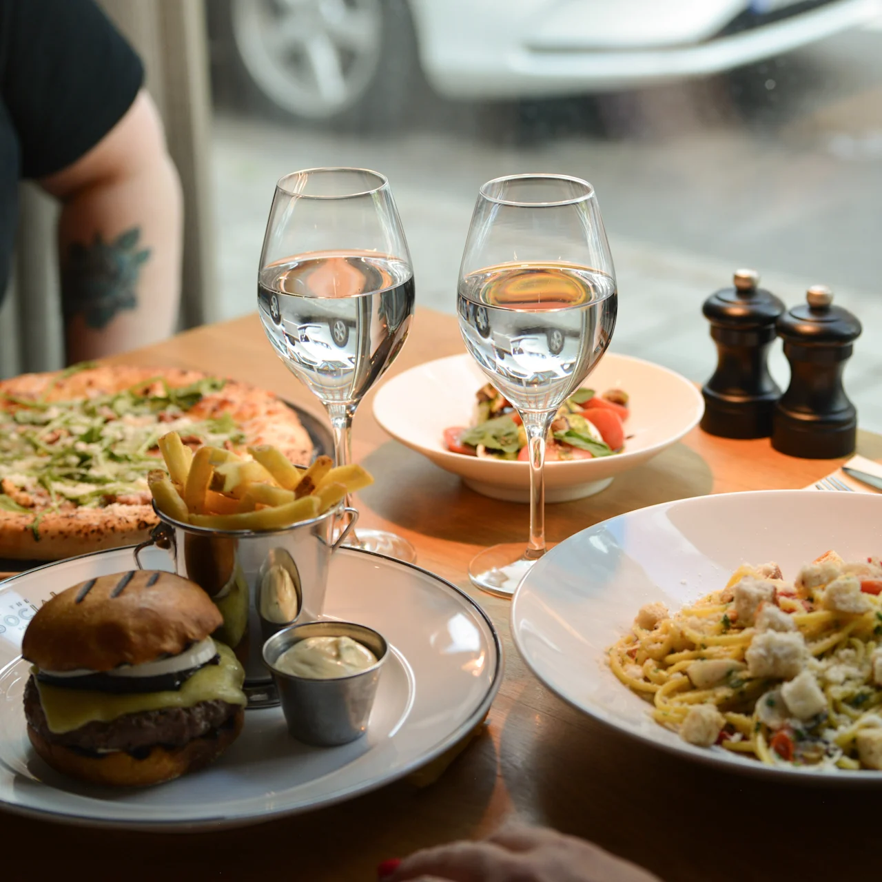Burger og pommes frites sidder på en tallerken med dyppesauce ved siden af et bord med pasta, pizza, salat og to glas vand, udsigt til en gade gennem et restaurantvindue.