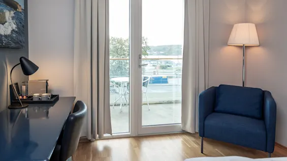 Et værelse med en blå lænestol ved siden af en tændt gulvlampe. Et skrivebord med en lampe og kopper står ved siden af. Glasdøre åbner ud til en altan med udsigt.