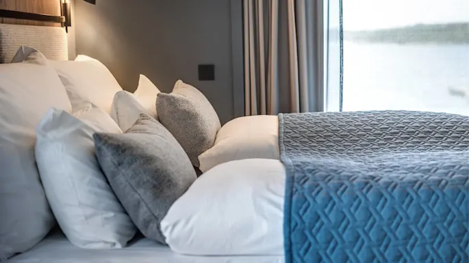 A neatly made bed has multiple white and grey pillows and a textured blue blanket in a softly lit room, next to a window with light curtains.