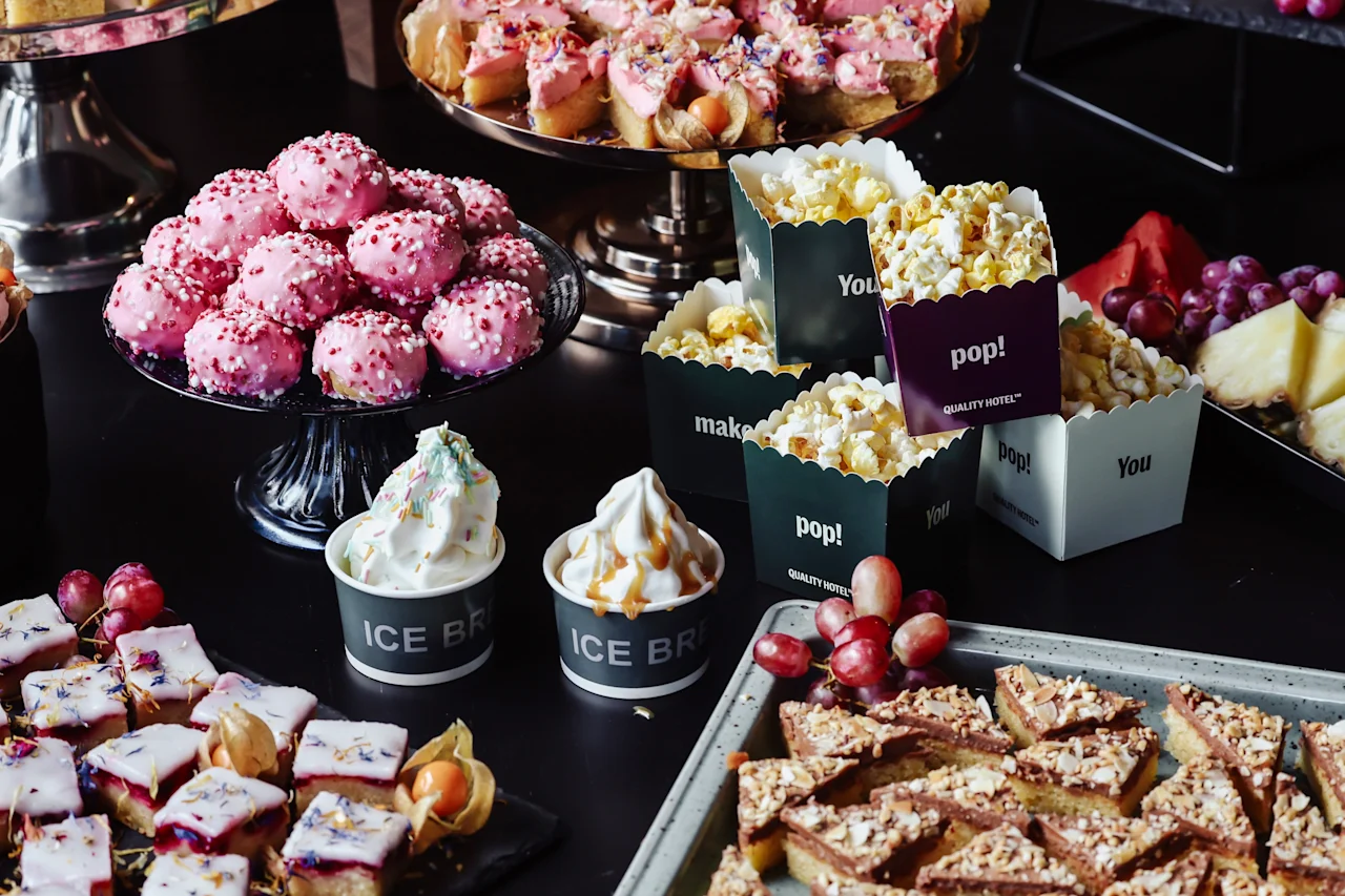 Pink pastries rest on a glass stand; next to them are popcorn in labeled containers, ice cream cups, and assorted fruits on a dark table. Containers read: "pop!", "ICE BRI", "make!", "You! QUALITY STREET".