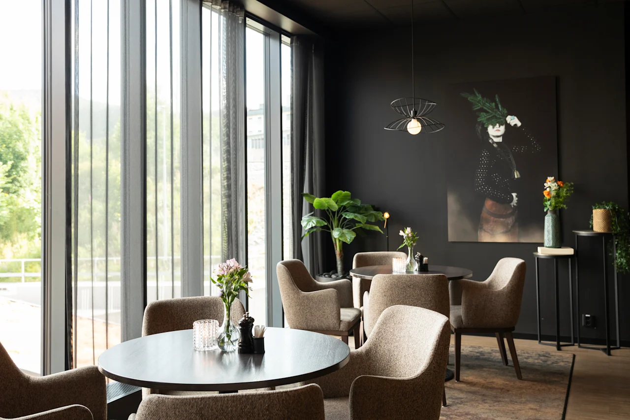 Et elegant og innbydende salongområde med komfortable sitteplasser, runde bord og store vinduer mot grøntområder.