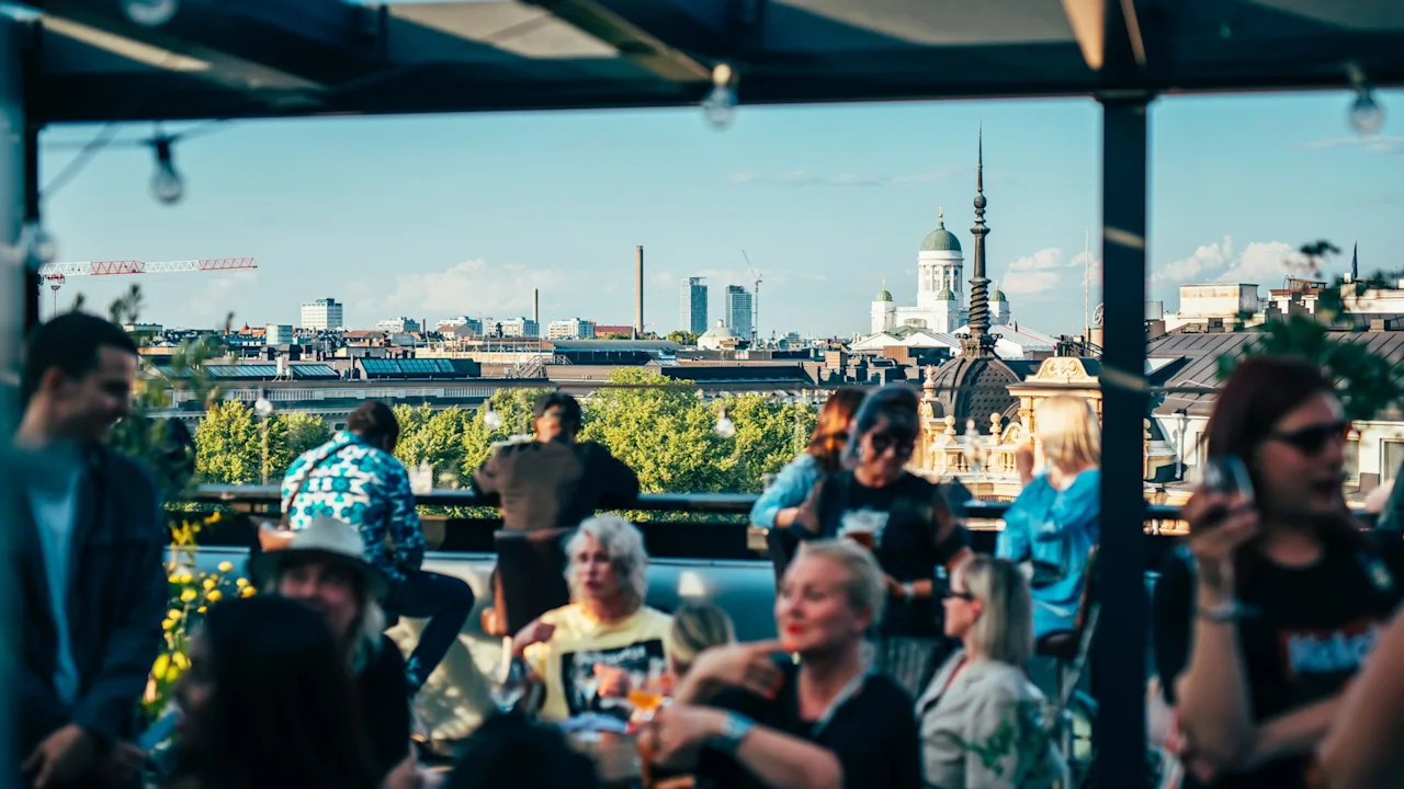 People are socializing on a rooftop terrace. They are sitting and standing with drinks while a cityscape, featuring buildings and a blue sky, provides the backdrop.