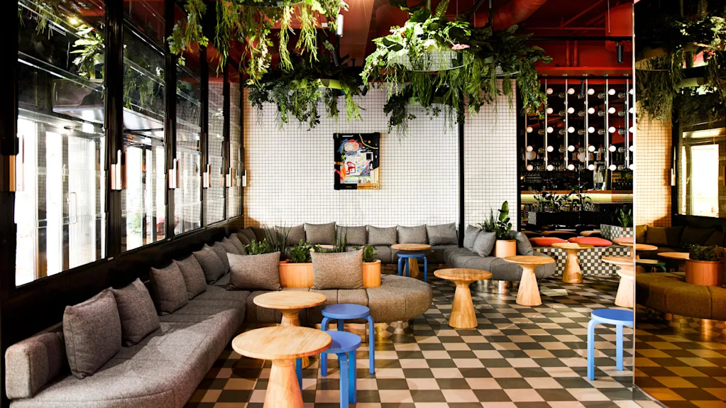 Gray couches form a semi-circle around wooden tables, with overhead green plants decorating the modern room featuring a checkered floor and large windows.